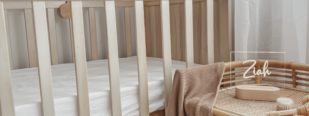 A cozy nursery setup featuring a wooden crib with a soft white mattress. Next to the crib is a wicker storage basket containing a beige blanket and baby essentials. A personalised Warm White Night Light from The Willow Corner, in the Smooth Script design, sits on the table, glowing softly with the name