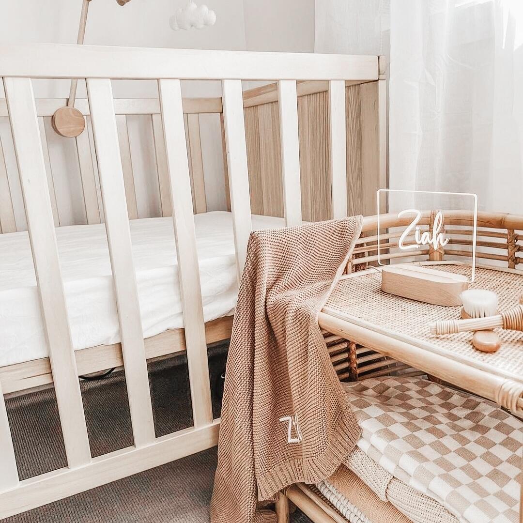 A cozy nursery setup featuring a wooden crib with a soft white mattress. Next to the crib is a wicker storage basket containing a beige blanket and baby essentials. A personalised Warm White Night Light from The Willow Corner, in the Smooth Script design, sits on the table, glowing softly with the name 